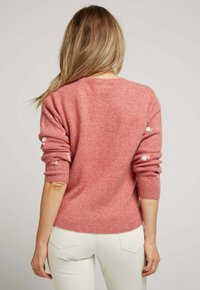 Pull en maille rose avec un col rond et des accents floraux brodés sur les manches. Le tissu semble doux et texturé.