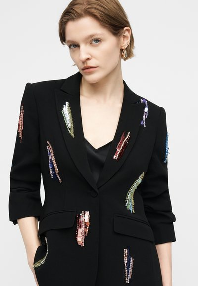 Γυναίκα με κοντά ανοιχτό καστανά μαλλιά που φοράει μαύρο σακάκι διακοσμημένο με πολύχρωμες παγιέτες και χρυσούς κρίκους σκουλαρίκια.