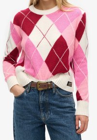 Donna che indossa un maglione lavorato a maglia argyle rosa, rosso e bianco, infilato in jeans blu con una cintura in pelle marrone, mano in tasca.