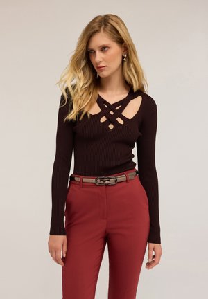 Haut à manches longues côtelé marron foncé avec un design en croix et des découpes aux épaules, associé à un pantalon rouge taille haute et une ceinture.