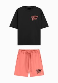 Zwarte T-shirt met korte mouwen en roze logo; gecombineerd met roze shorts met een elastische tailleband en zwart logo aan de zoom. Katoenen stof.