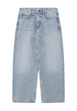 Personne portant un jean large bleu clair, un haut sans manches jaune moutarde à boutons, et des sandales marron cloutées, les mains dans les poches.