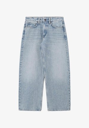 Personne portant un jean large bleu clair, un haut sans manches jaune moutarde à boutons, et des sandales marron cloutées, les mains dans les poches.