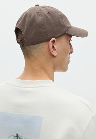 Mand iført en brun baseballkasket og en hvid sweatshirt med en rektangulær grafik på ryggen, vendt mod højre foran en ensfarvet baggrund.