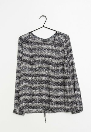 Blouse à manches longues à motif noir et blanc, col rond, ourlet froncé avec cordon, suspendue sur un cintre noir sur fond blanc.