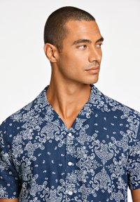 Camisa de manga corta hecha de tela azul con un patrón de paisley blanco, diseño de botones y cuello de campamento. Apariencia de lienzo texturizado.