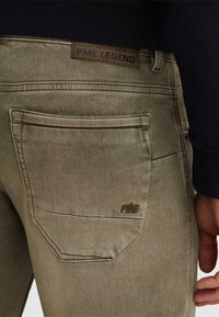 Vista trasera de unos jeans color caqui con un bolsillo que presenta un bordado con las letras "PME" y una etiqueta de cuero encima de la pretina que dice "PME LEGEND".