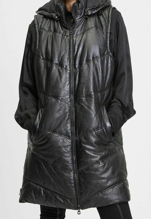 Personne portant une longue veste sans manches en cuir noir matelassée avec une capuche et une fermeture éclair à l’avant, mains dans les poches latérales, par-dessus un tee-shirt noir à manches longues.