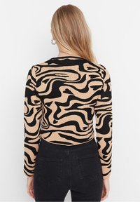 Beige gestricktes Oberteil mit schwarz-abstractem Wirbmuster, mit langen Ärmeln und einer figurnahen Silhouette. Kombiniert mit schwarzen Jeans.