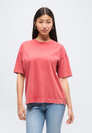 Ung kvinde med langt sort hår iført en falmet rød GAP t-shirt og lyseblå jeans, stående mod en ensfarvet hvid baggrund.