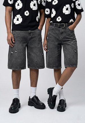Deux personnes portant des hauts à motifs floraux noirs et blancs, des shorts en jean noirs déchirés, des chaussettes blanches et des chaussures noires épaisses, debout côte à côte.