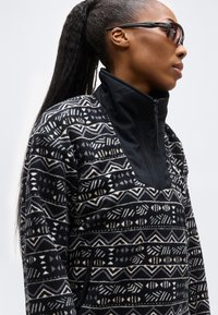 Γυναίκα με μακριά πλεξούδα, που φοράει σκούρο φλις μπουφάν με φερμουάρ και γυαλιά ηλίου που τυλίγουν γύρω, κοιτάζει προς τα δεξιά, σε απλό φόντο.