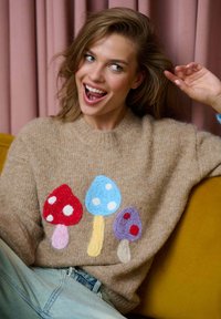 Beżowy sweter o puszystej teksturze, z trzema filcowymi wzorami grzybów w kolorze czerwonym, niebieskim i purpurowym z kropkami. Niebieskie tło.