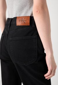 Persona che indossa jeans neri con una toppa in pelle marrone Calvin Klein sulla vita, indossando una maglietta grigia chiara a costine, con un braccio visibile che mostra una cicatrice.