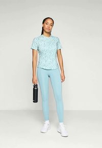 Naine seisab helesinisest mustriga T-särgist ja legginsitest, valgetes tossudes, hoides musta veepudelit ühtlase heleda taustaga.