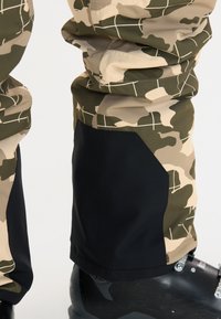 Camo broek in groen, bruin en beige met zwarte accenten; heeft een duurzame stof en verstevigde onderbenen. Laarzen zichtbaar aan de onderkant.