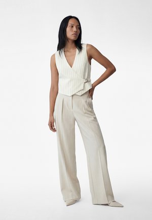 Mujer de pie con la mano en la cadera, vistiendo un chaleco cruzado sin mangas de rayas crema y pantalones de pierna ancha a juego, con zapatos puntiagudos.