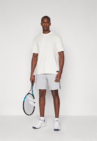 Mäns ljusgrå shorts och off-white kortärmad t-shirt, hållande en tennisracket. Tyget verkar jämnt med moderat passform och utan mönster.