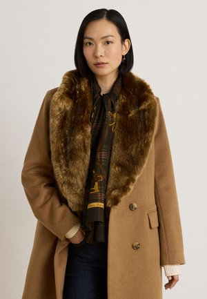 Femme debout à l'intérieur portant un long manteau camel avec un grand col en fourrure, un jean foncé, des bottes marron à talons et tenant un sac à main rond marron.