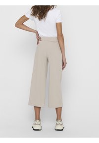 Frau steht mit der Hand auf der Hüfte, trägt ein weißes kurzärmeliges Oberteil, beigen weit geschnittenen, kniehohen Hosen und weiße Turnschuhe, vor einem einfarbigen Hintergrund.