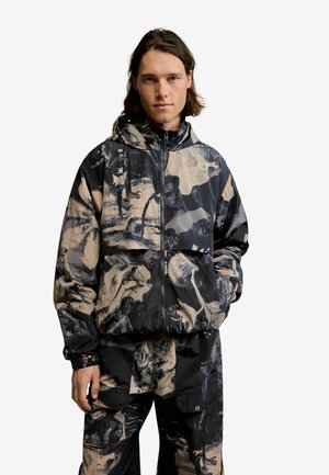 Junger Mann mit schulterlangem Haar, der eine schwarze und beige Kapuzenjacke mit abstraktem Muster und passende Hosen trägt, die Hände in den Taschen.