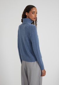 Maglione blu in lana con collo alto e orlo ribattuto, caratterizzato da una texture morbida e una vestibilità comoda, abbinato a pantaloni grigi chiari.