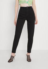 Zwarte high-waisted jeans met een getailleerd silhouet, vijf zakken en een knoopsluiting, gecombineerd met zwarte geperforeerde hakmuilen.