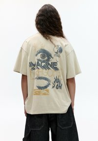 Beige oversize tričko s grafickým potlačou na zadnej strane, ktorá zobrazuje oko, plamene a text "Imagine Dragons" v tmavomodrej a zlatej farbe.