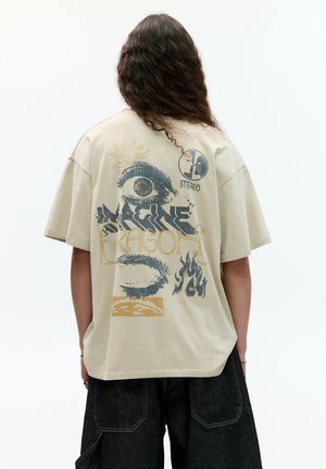 Beżowy, oversize t-shirt z grafiką na plecach przedstawiającą oko, płomienie oraz tekst "Imagine Dragons" w ciemnoniebieskich i złotych akcentach.