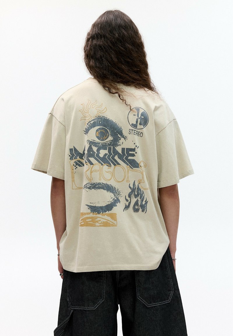 Beige oversize tričko s grafickým potlačou na zadnej strane, ktorá zobrazuje oko, plamene a text "Imagine Dragons" v tmavomodrej a zlatej farbe.