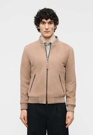 Jeune homme aux cheveux bouclés portant une veste bomber beige zippée par-dessus une chemise gris clair et un pantalon noir, debout devant un fond uni.