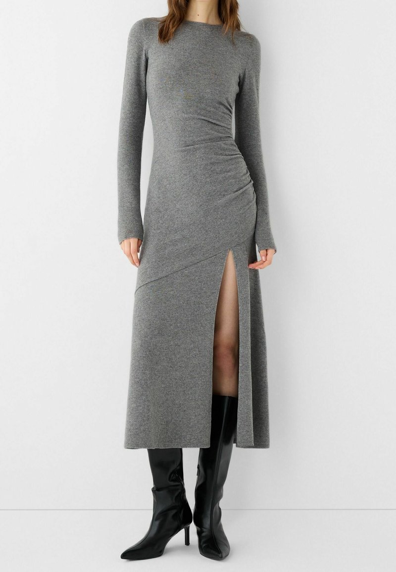 Graues Longsleeve-Kleid aus weichem Stoff mit seitlichem Schlitz und gerafftem Detail an der Taille, kombiniert mit schwarzen kniehohen Stiefeln mit Stilettoabsatz.