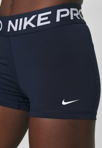 Tummansininen urheilushortsit, joissa on valkoinen "NIKE PRO" vyötärö ja pieni valkoinen Nike-logo vasemmassa alakulmassa. Sujuva, joustava kangas, tiukka muotoilu.
