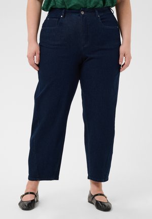 Femme portant un jean bleu foncé taille haute, coupe droite, un haut vert et des chaussures Mary Jane noires, debout avec les mains le long du corps sur un fond uni.