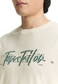 Roomwitte katoenen T-shirt met een ronde hals, voorzien van groene tekstgrafieken en zwarte accenten. Zachte textuur, met bedrukte details en een ontspannen pasvorm.