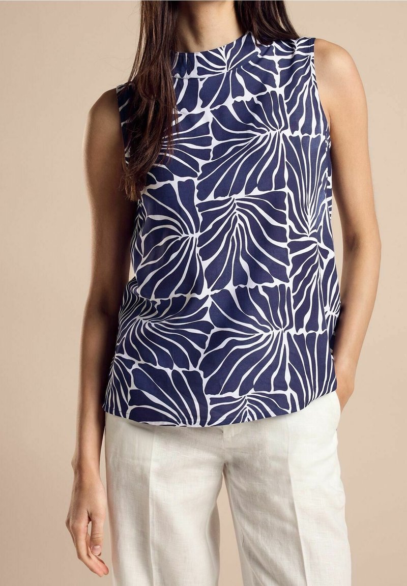 Femme portant un haut bleu marine sans manches avec des motifs abstraits de feuilles blanches et un pantalon blanc sur un fond beige uni.