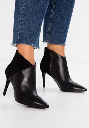 Bottes en cuir noires à bout pointu et talons aiguilles, portées avec un jean bleu à ourlet effiloché, sur fond blanc.