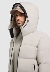 Polstret beige jakke med en sort-belagt hætte, frontlynlås og sidelommer. Modellen bærer en beige cap med et logo. Glat, isoleret tekstur.