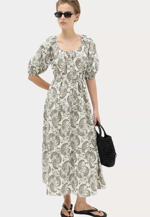 Vrouw draagt een witte midi-jurk met paisley patroon en pofmouwen, zwarte zonnebril, houdt een zwarte gevlochten handtas vast en zwarte sandalen.