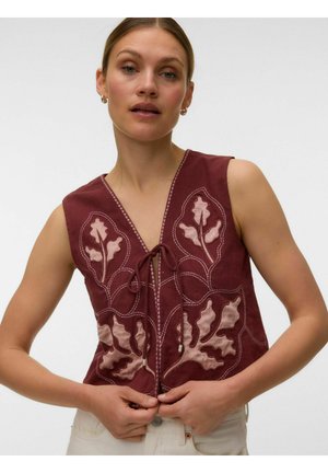 Vrouw draagt een mouwloos kastanjebruin vest met roze bladborduursel, strik vooraan, gecombineerd met een witte broek, neutrale achtergrond.