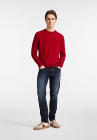 Crvena sweatshirt s okruglim izrezom, nosi se s tamnoplavim trapericama i bež tenisicama s smeškim i krem akcentima. Model stoji s rukama u džepovima.