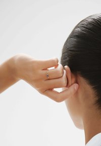 Anillo de oro con dos pequeñas piedras azules, llevado en un dedo, mientras una mano toca una oreja. El cabello está peinado hacia atrás, mostrando una textura suave.
