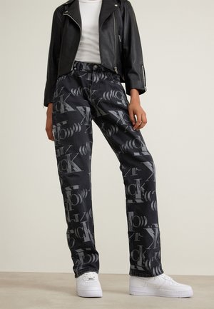 Osoba nosící černou koženou bundu, bílou košili, černé džíny se šedými abstraktními písmeny a bílé tenisky stojí před neutrálním pozadím.