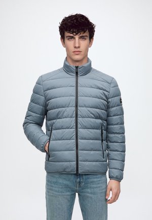 Jeune homme portant une veste matelassée bleu clair avec fermeture éclair frontale et poches, se tenant devant un fond clair uni.