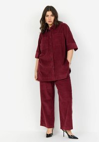 Set composto da camicia e pantaloni in corduroy bordeaux. La camicia presenta maniche corte, due tasche sul petto e una chiusura a bottoni. I pantaloni sono di stile a gamba larga.