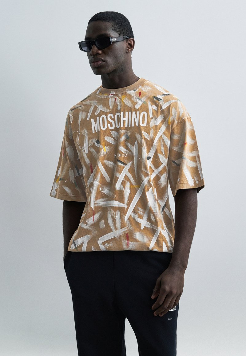 Άνδρας που φοράει γυαλιά ηλίου, ένα καφέ πουκάμισο Moschino με λευκές πινελιές που μοιάζουν με χρώμα και μαύρο παντελόνι, με τα χέρια στις τσέπες, μπροστά σε απλό φόντο.