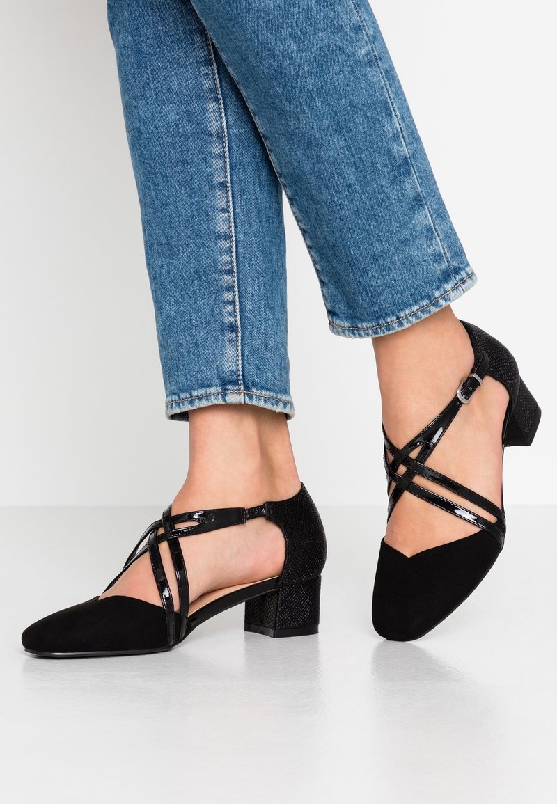 Femme portant un jean bleu et des chaussures noires à talons bas avec des brides croisées en cuir verni et une boucle, posée sur une surface blanche.