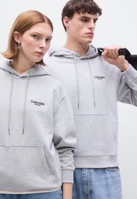 Γκρι φούτερ με κουκούλα, μπροστινές τσέπες και κεντημένο λογότυπο "Calvin Klein Jeans". Τα μοντέλα φορούν τζιν παντελόνια με άνετη εφαρμογή.