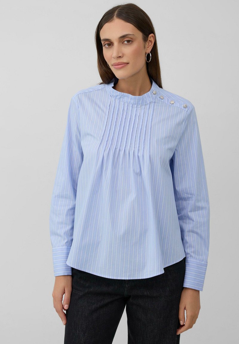 Blusa azzurra a maniche lunghe con strisce bianche verticali, pieghe sul davanti e bottoni decorativi lungo il colletto. Tessuto dalla consistenza liscia.