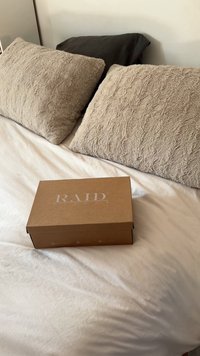 Brązowe pudełko kartonowe z napisem "RAID LONDON" na białym łóżku, w tle teksturowane beżowe i ciemne poduszki.